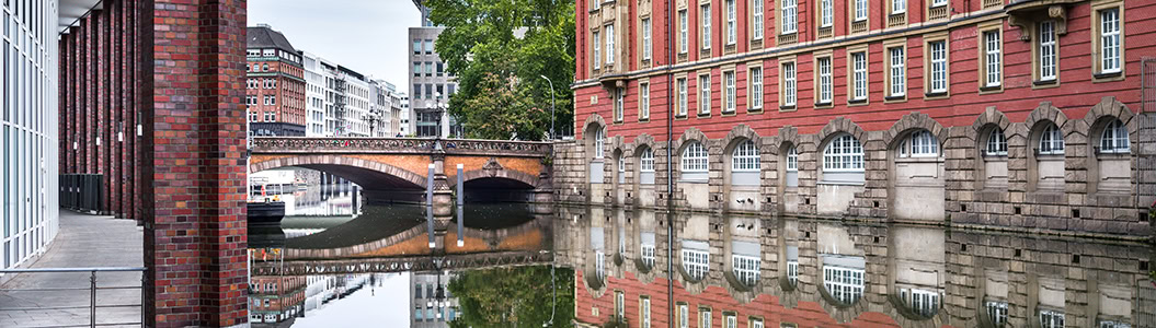 Breiter Kanal mit rotem Backsteingebäude, Brücke und üppigem Grün im Hintergrund.